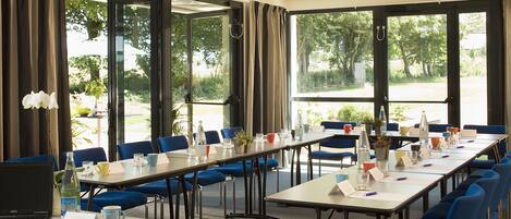 Un salón de conferencias con mesas largas, sillas azules y ventanas amplias con cortinas.