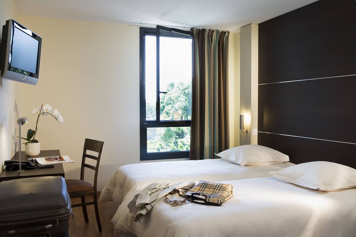 A hotel room with a bed, a desk with a lamp, a chair, a suitcase, and a window with curtains.
