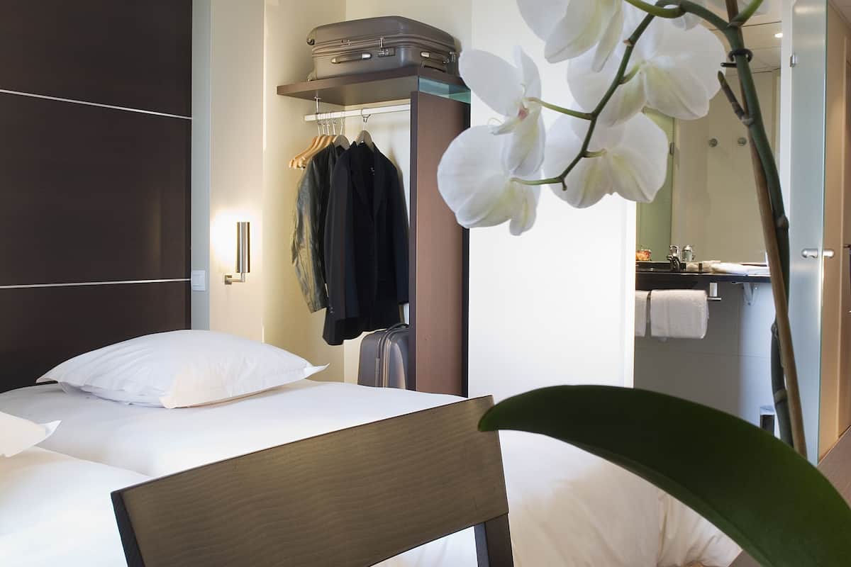 A hotel room with a bed, a chair, a potted orchid, a hanging coat rack, and a bathroom with a towel rack.