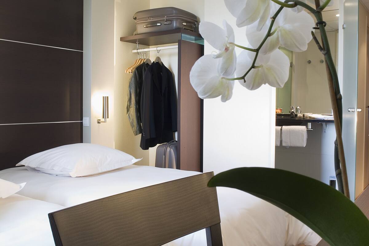 A hotel room with a bed, a chair, a potted orchid, a hanging coat rack, and a bathroom visible through an open door.