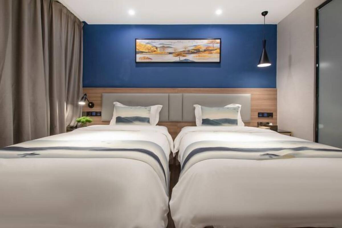 A hotel room with two beds, a blue accent wall, a painting, and a pendant light.