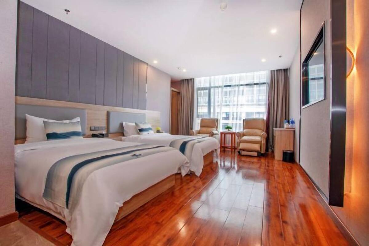 A hotel room with two beds, a small table, a chair, and a large window.