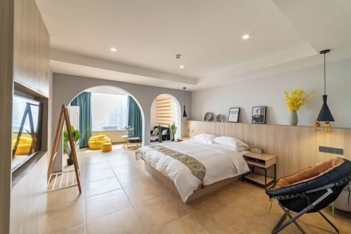 A modern bedroom with a large bed, a sitting area with a yellow chair, and a view of the city through large windows.