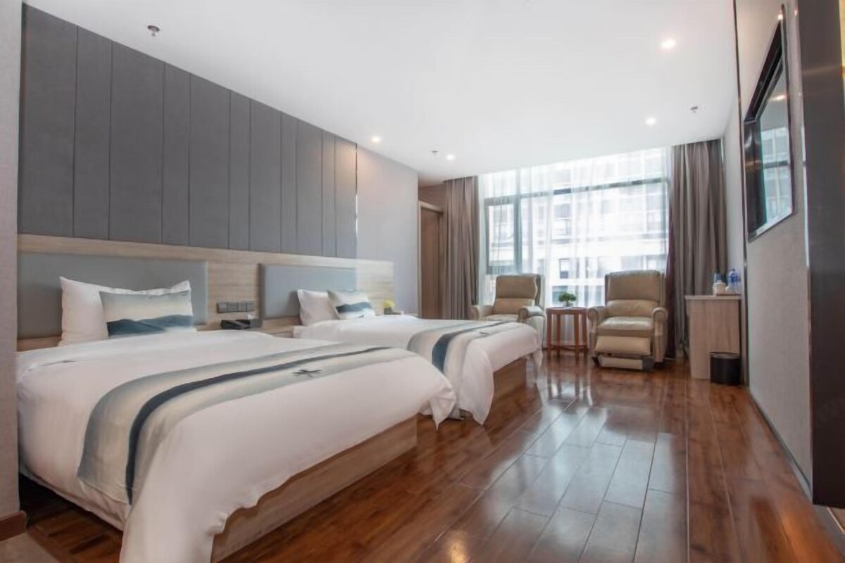 Habitación de hotel con dos camas, un área de descanso con sillas, una mesita y un ventanal que ofrece vistas de la ciudad.
