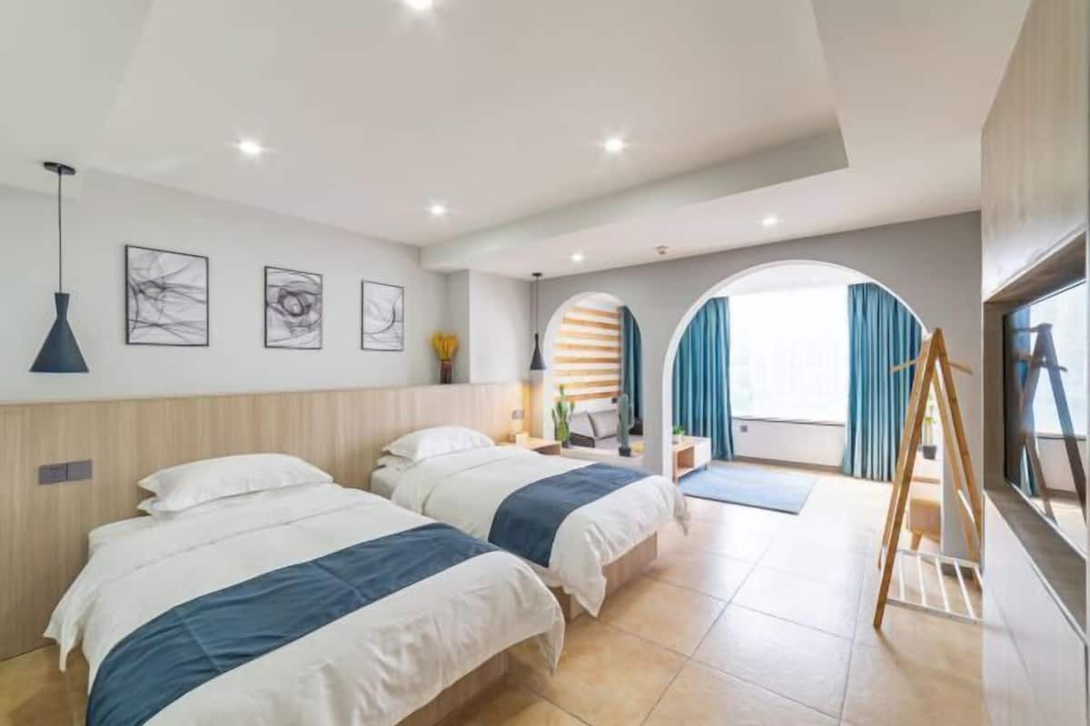 A hotel room with two beds, a standing desk, a large window with curtains, and artwork on the walls.