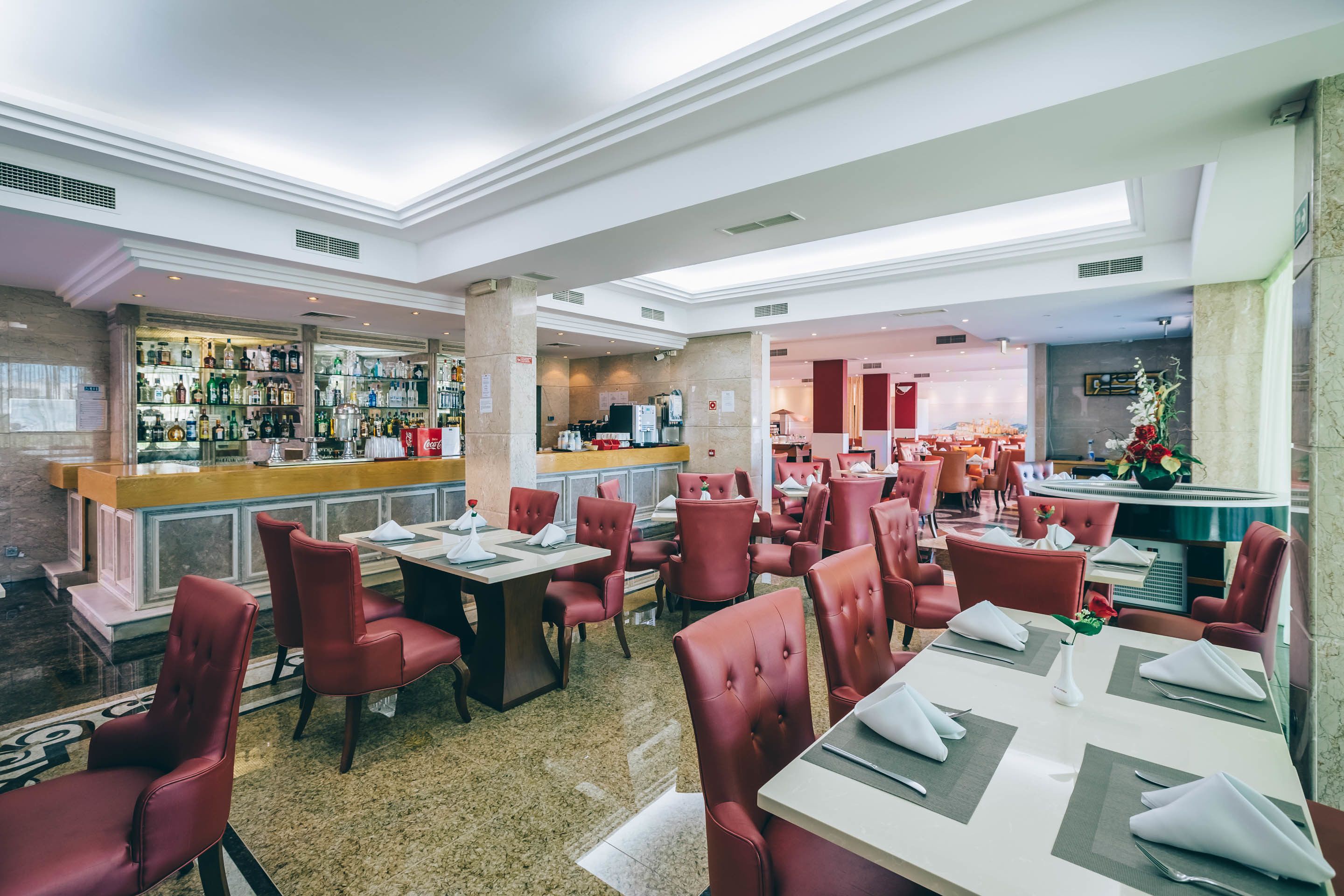 A restaurant with red upholstered chairs, neatly set tables, a bar area with various bottles, and a modern interior design.