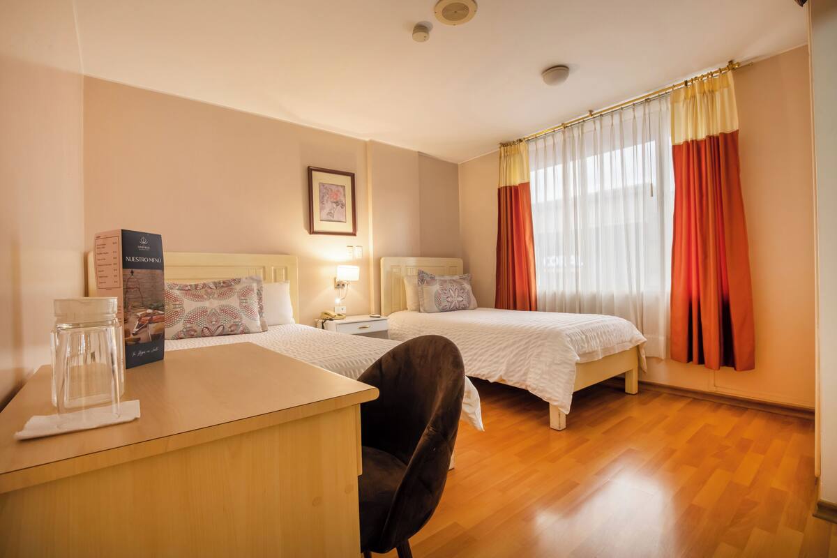 A hotel room with two beds, a desk, and a chair. There is a framed picture on the wall and a window with curtains.