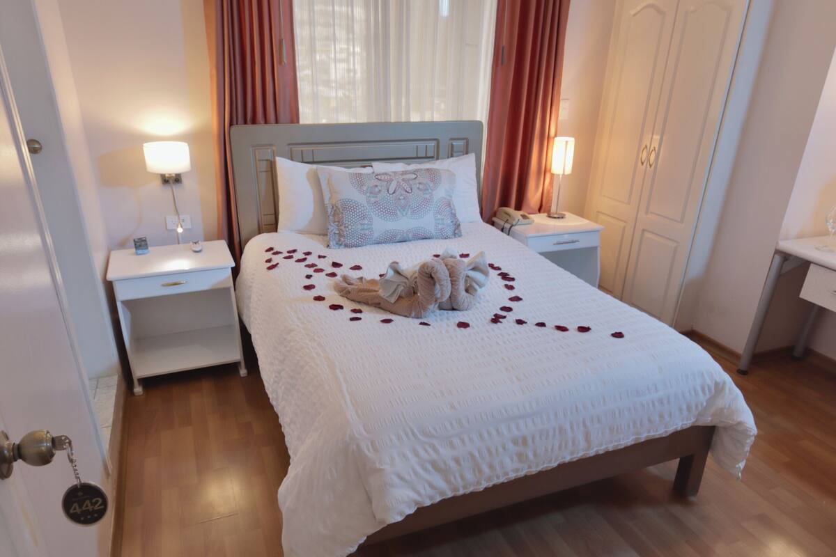 A neatly made bed with a white duvet, a bedside table with a lamp, and a wardrobe in a well-lit room with wooden flooring.