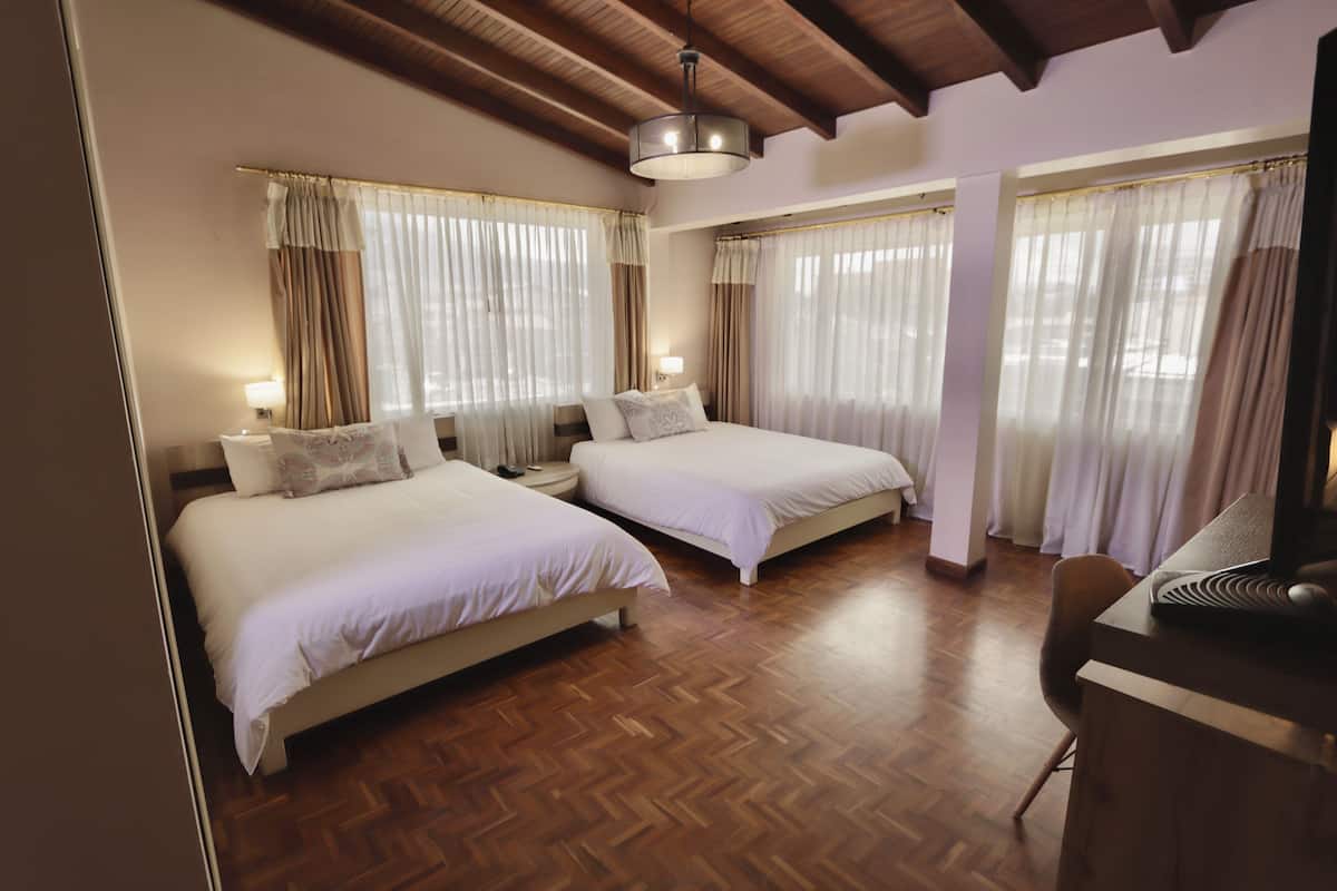 A hotel room with two beds, wooden flooring, and large windows with curtains.