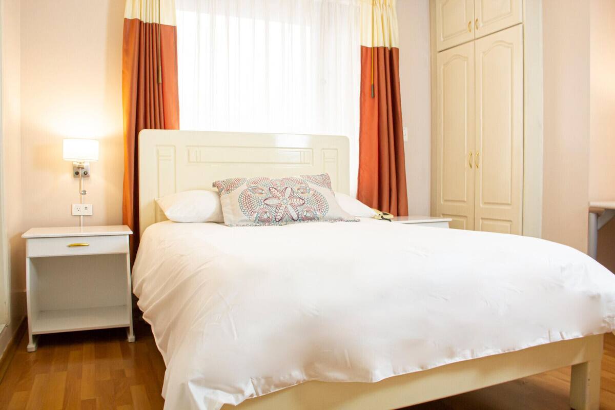 A neatly made bed with white bedding and a patterned pillow, a bedside table with a lamp, and a wardrobe in a well-lit room.