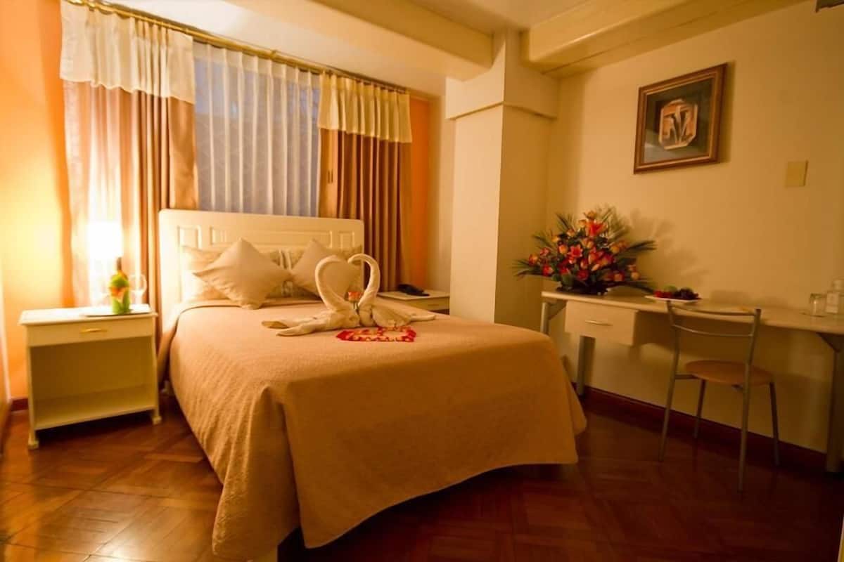 A hotel room with a bed, a desk, a chair, a nightstand, and a painting on the wall.
