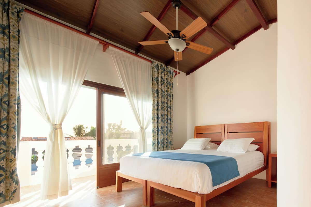 A bedroom with a wooden bed, white bedding, a ceiling fan, and large windows with curtains.