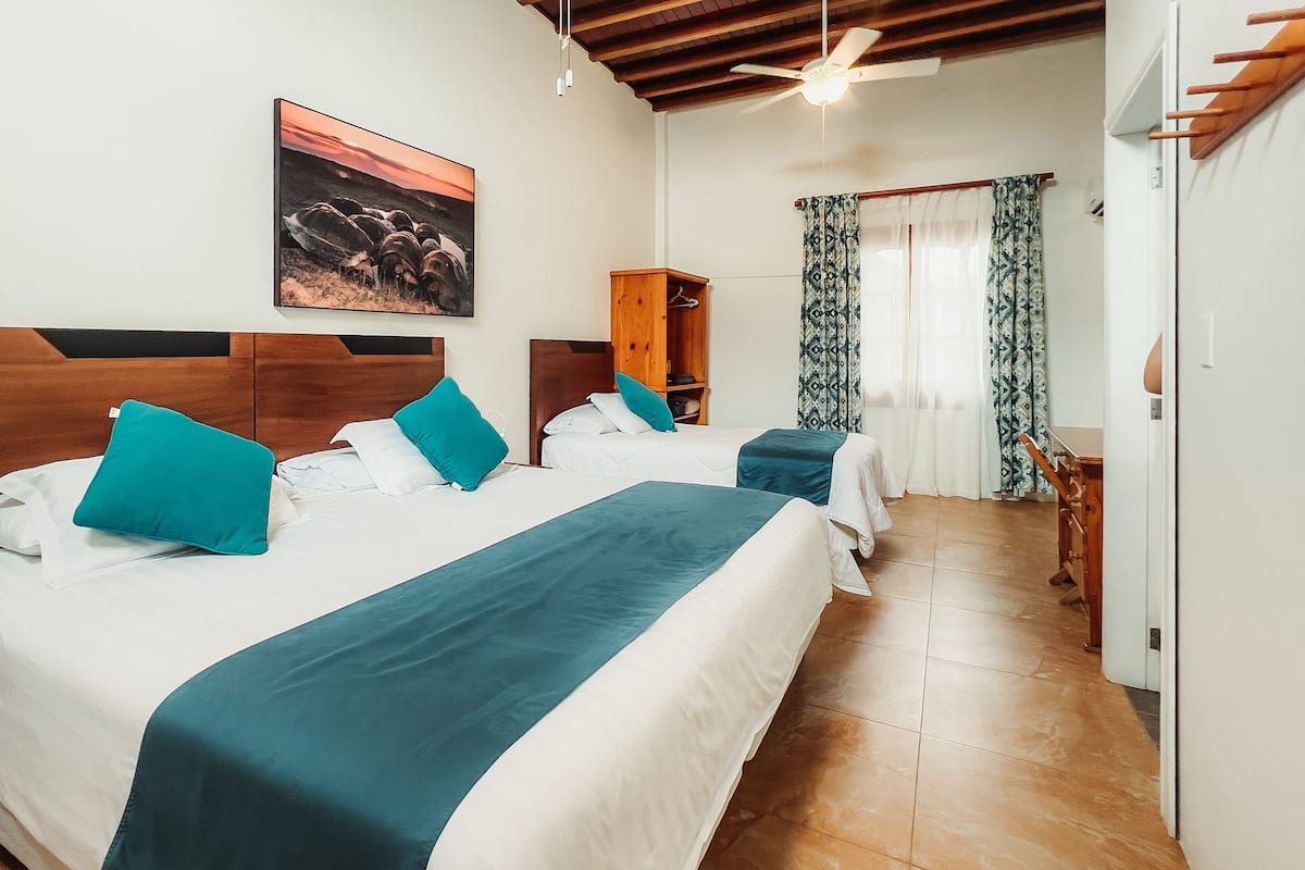 A hotel room with two beds, a wooden headboard, a ceiling fan, a painting, and a window with patterned curtains.
