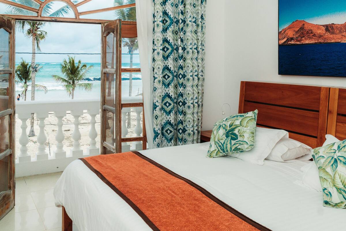 A bedroom with a bed, pillows, a view of the beach, and a painting of a mountain.