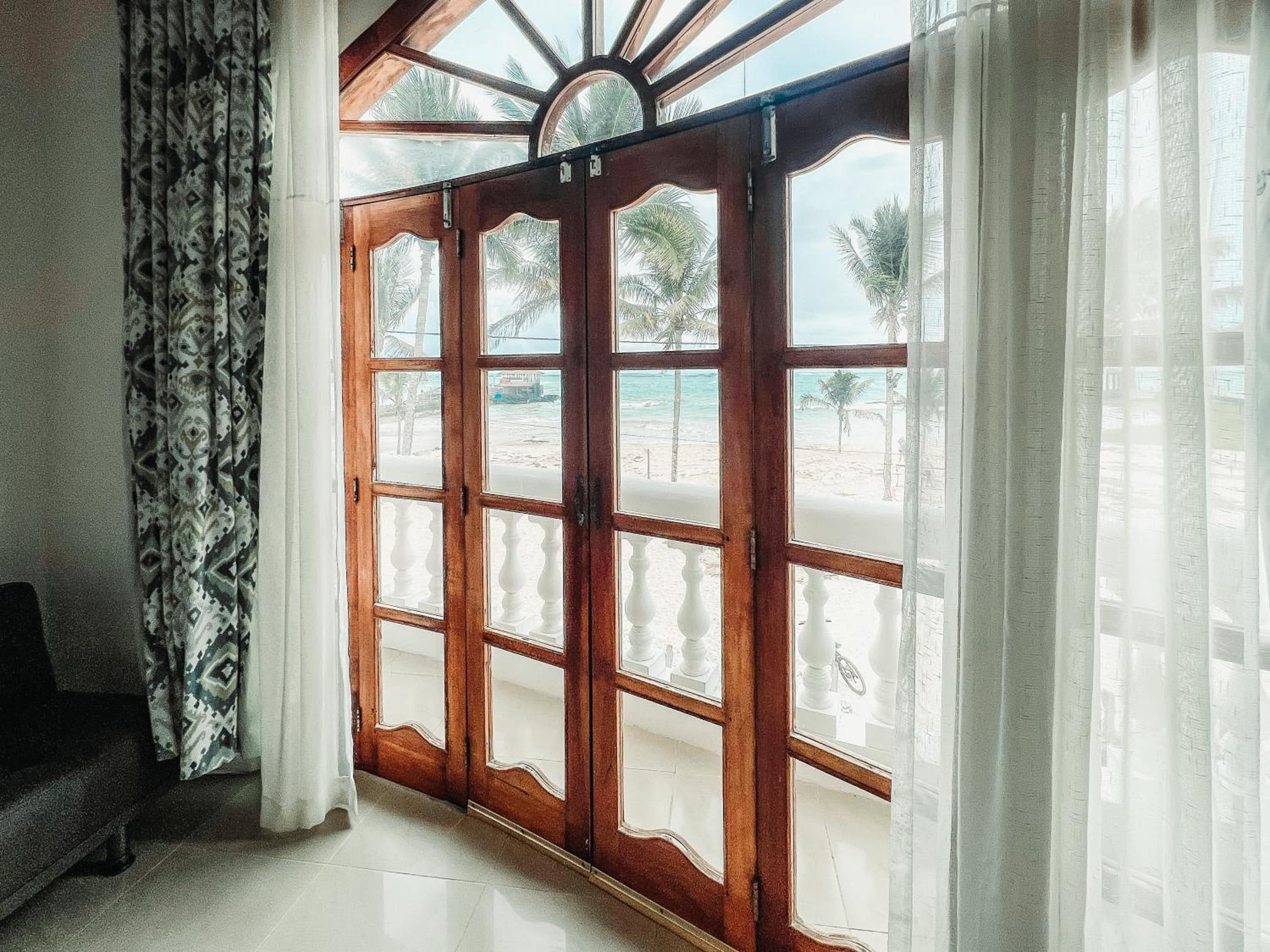 A room with a large wooden door leading to a beach view, a patterned curtain, and a sofa.
