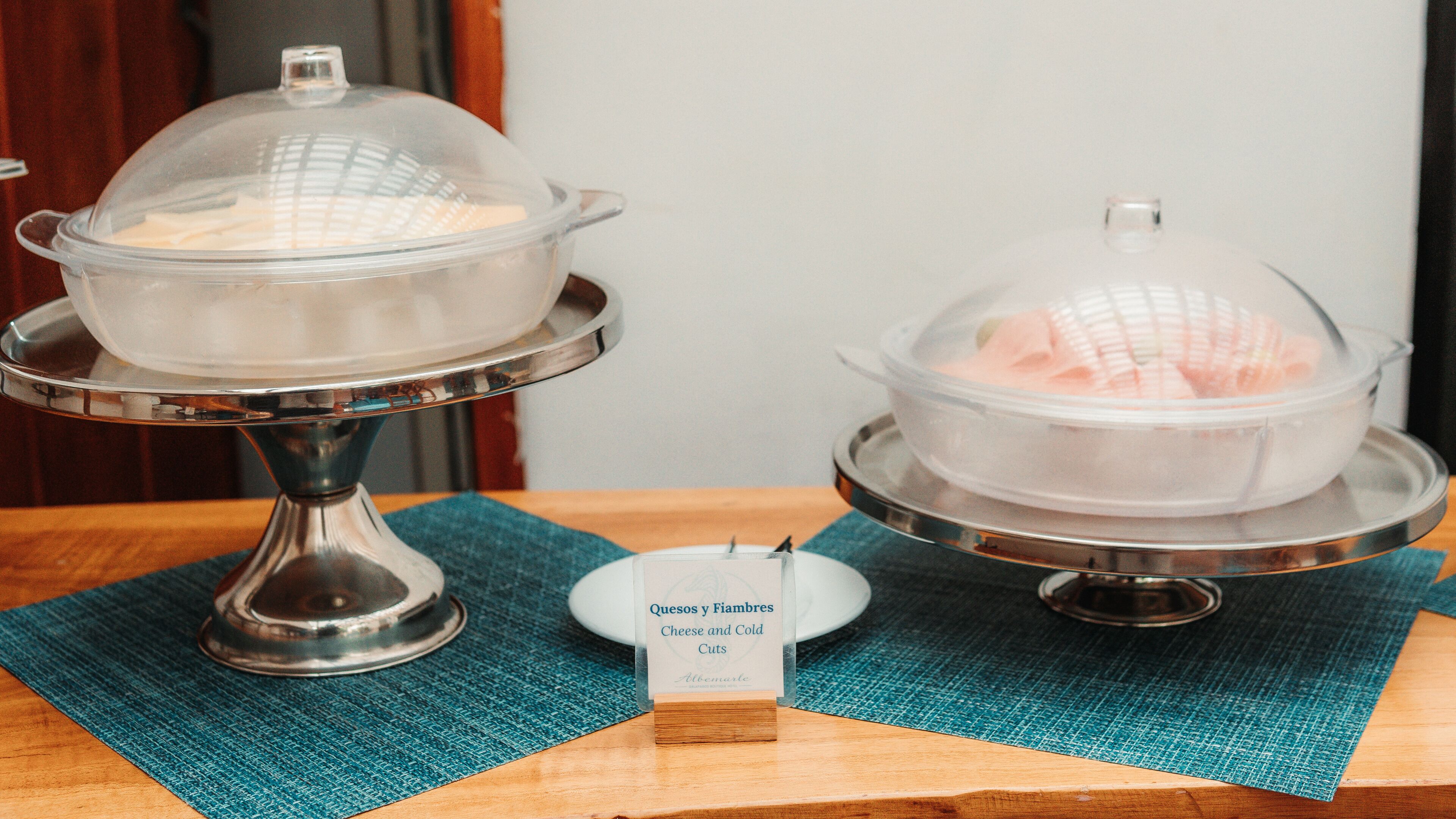 Two covered food dispensers labeled 'Quesos y Fiambres' on a wooden table with a blue placemat.