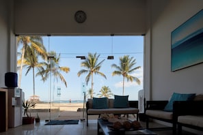 A living room with a large glass door leading to a beach view, featuring palm trees and a clear sky. - Hotel Albemarle Galapagos Beachfront (Puerto Villamil)