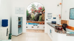 A reception area with a glass door leading to a pool area, a refrigerator stocked with beverages, and a sign indicating room numbers. - Hotel Albemarle Galapagos Beachfront (Puerto Villamil)