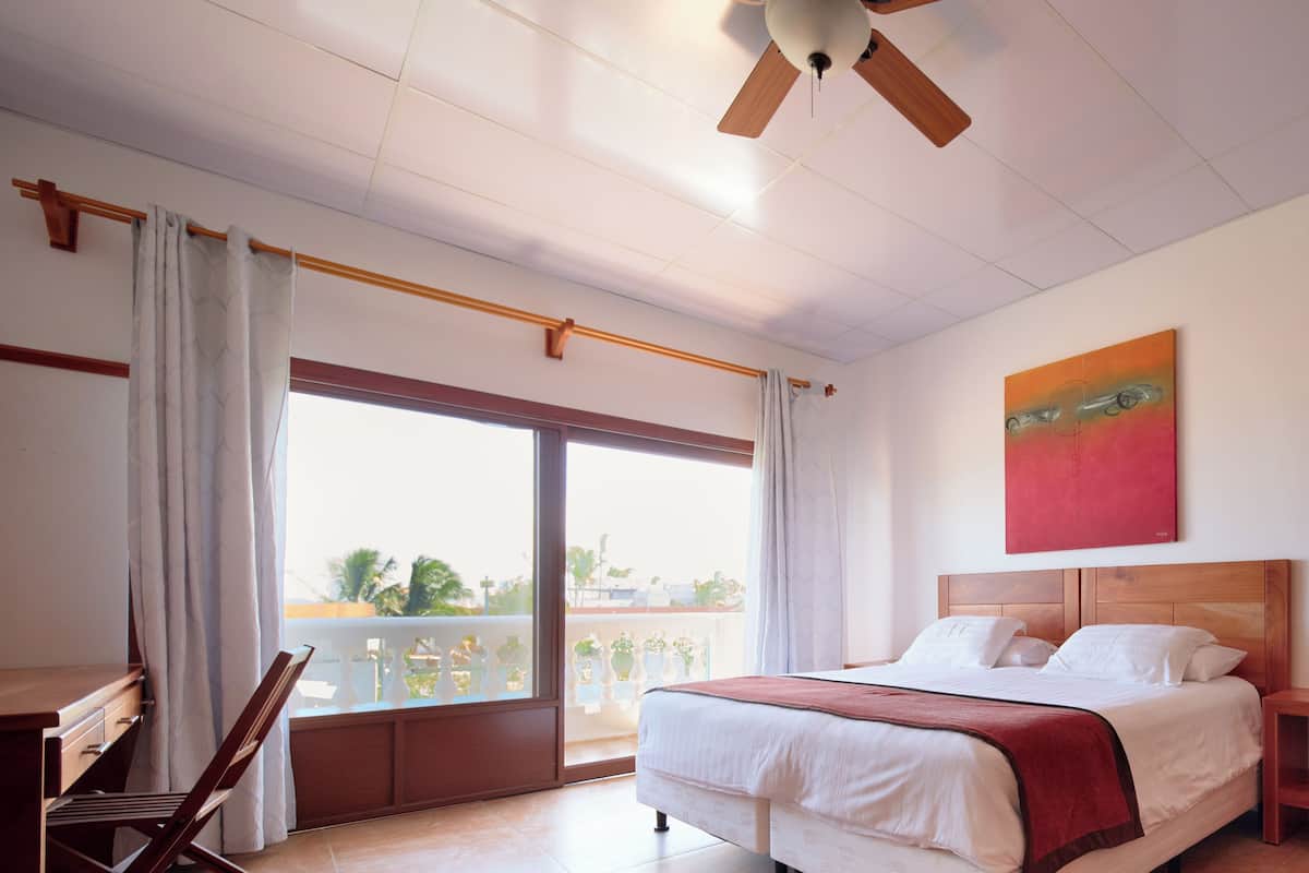 A hotel room with a bed, a desk, a chair, a balcony with a view, and a ceiling fan.