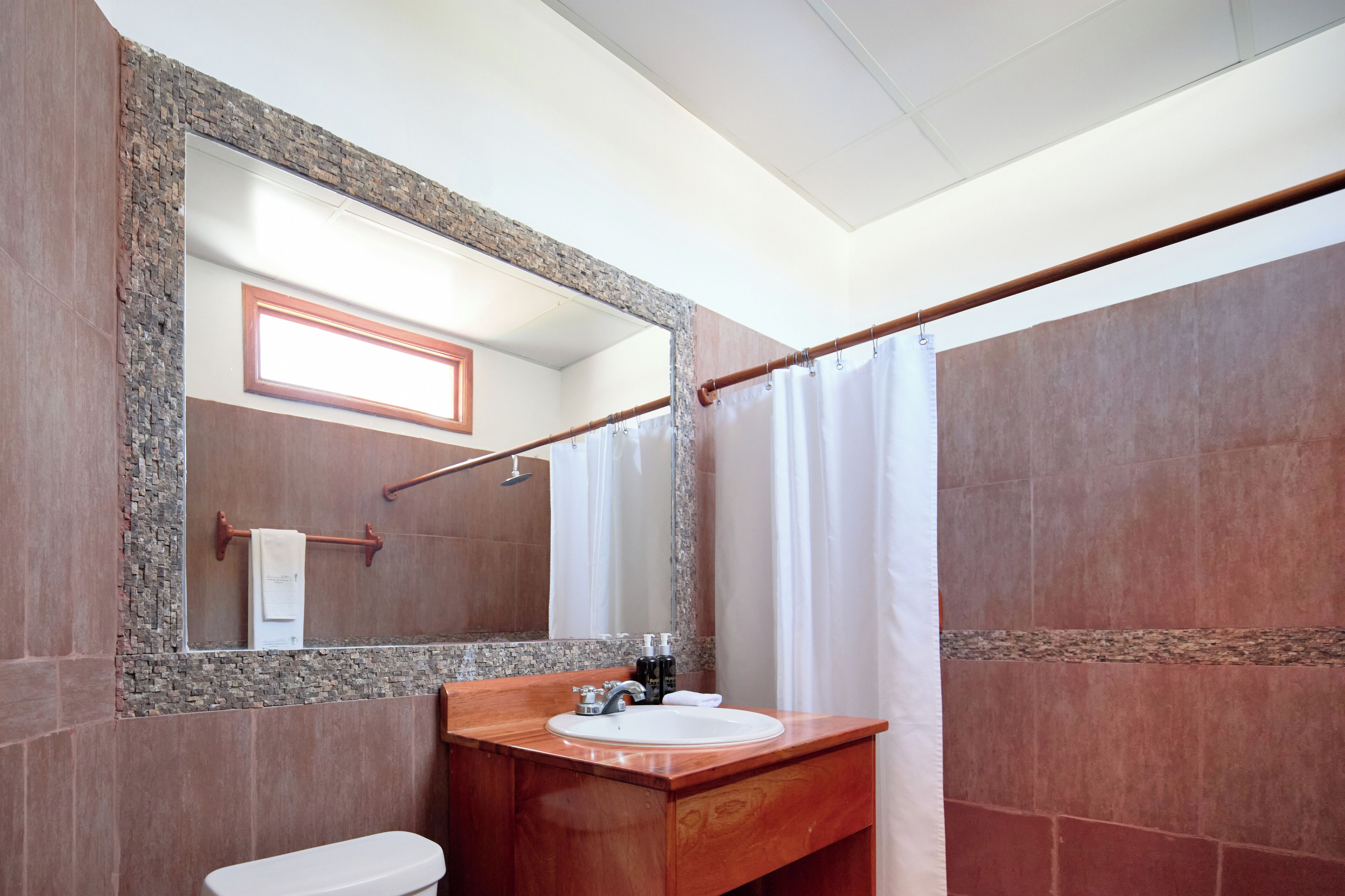 Un baño con tocador de madera, un lavamanos blanco, un espejo con marco de piedra y una cortina de ducha.