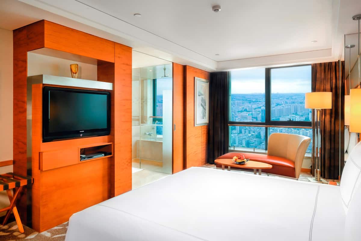Una habitación de hotel moderna con una cama grande, un televisor de pantalla plana, una mesita con fruta, una zona de estar y vistas a la ciudad a través de amplios ventanales.