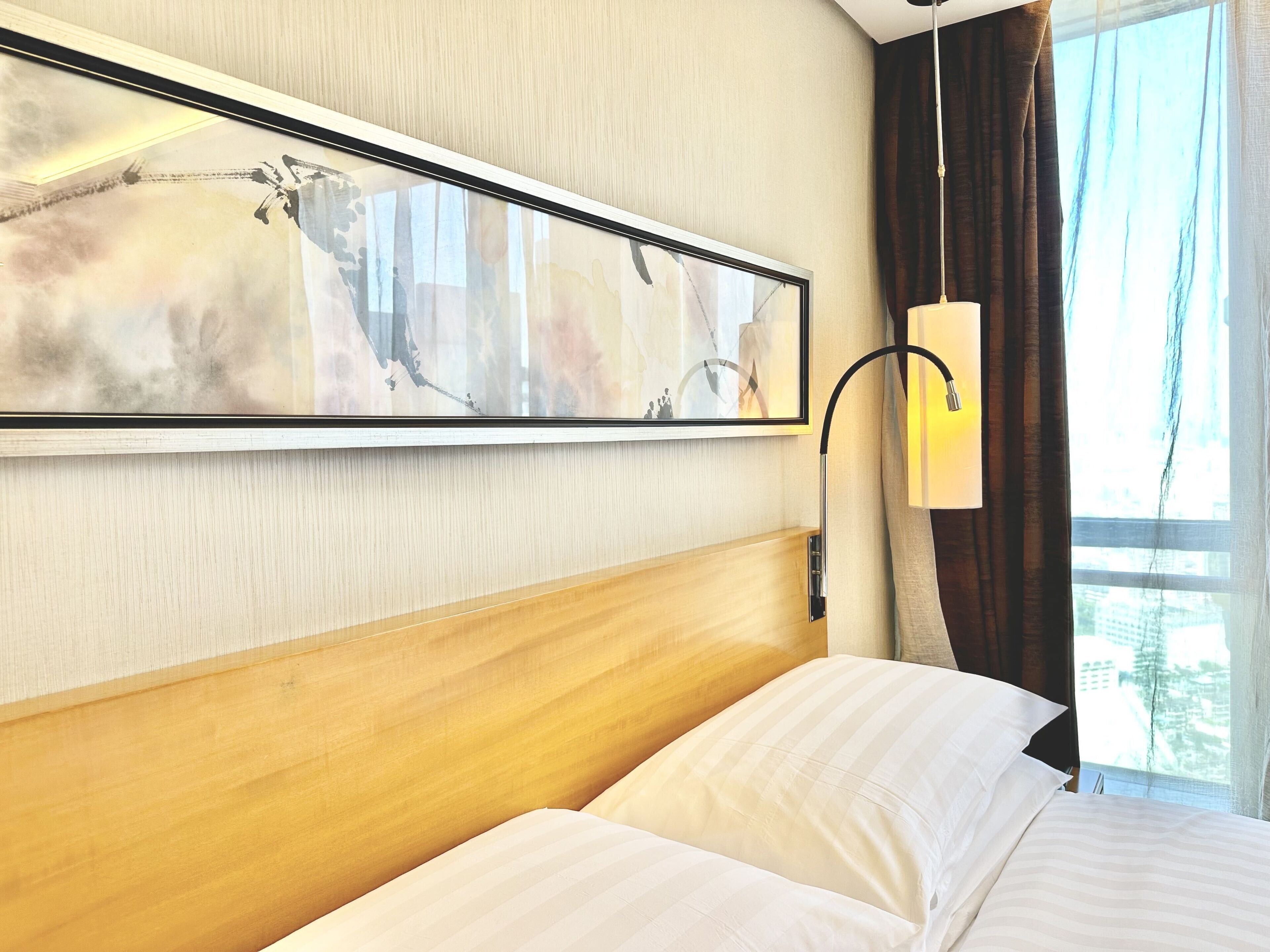 Habitación de hotel con cama, cabecero, una obra de arte enmarcada, lámpara y ventana con cortinas.