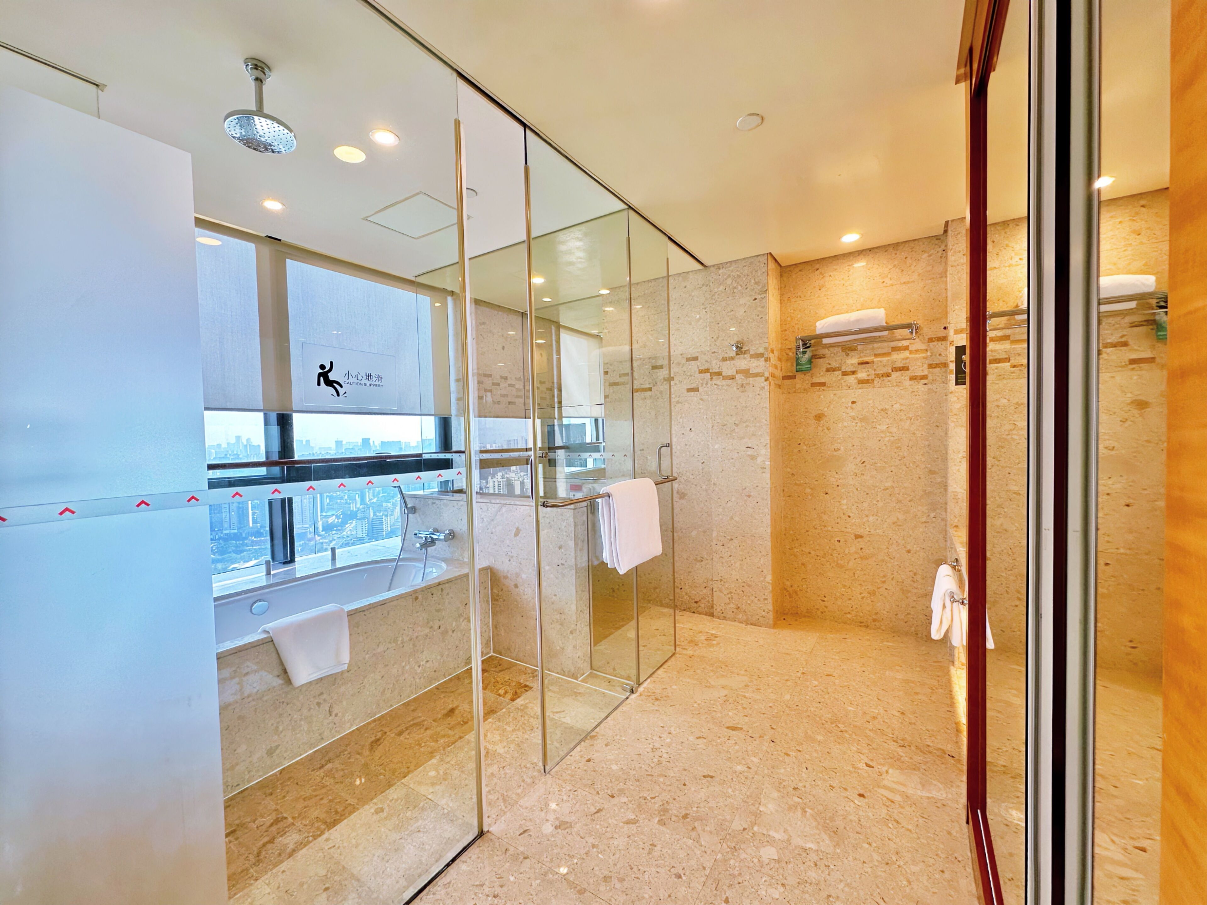 A modern bathroom with a glass shower enclosure, a sink with a towel rack, and a view of the city through a large window.