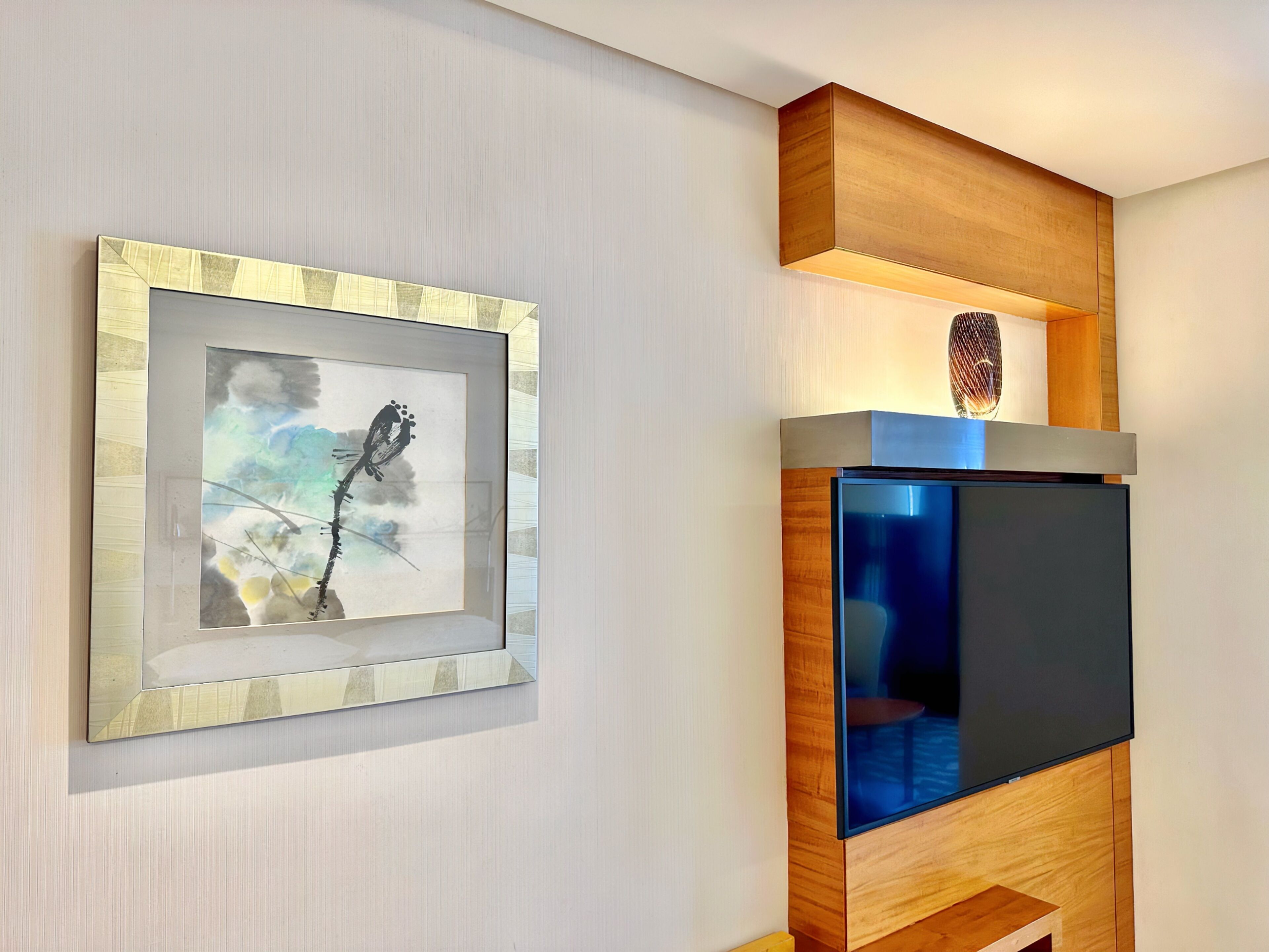 A framed artwork on a wall, a television on a wooden stand, and a shelf with a decorative item.