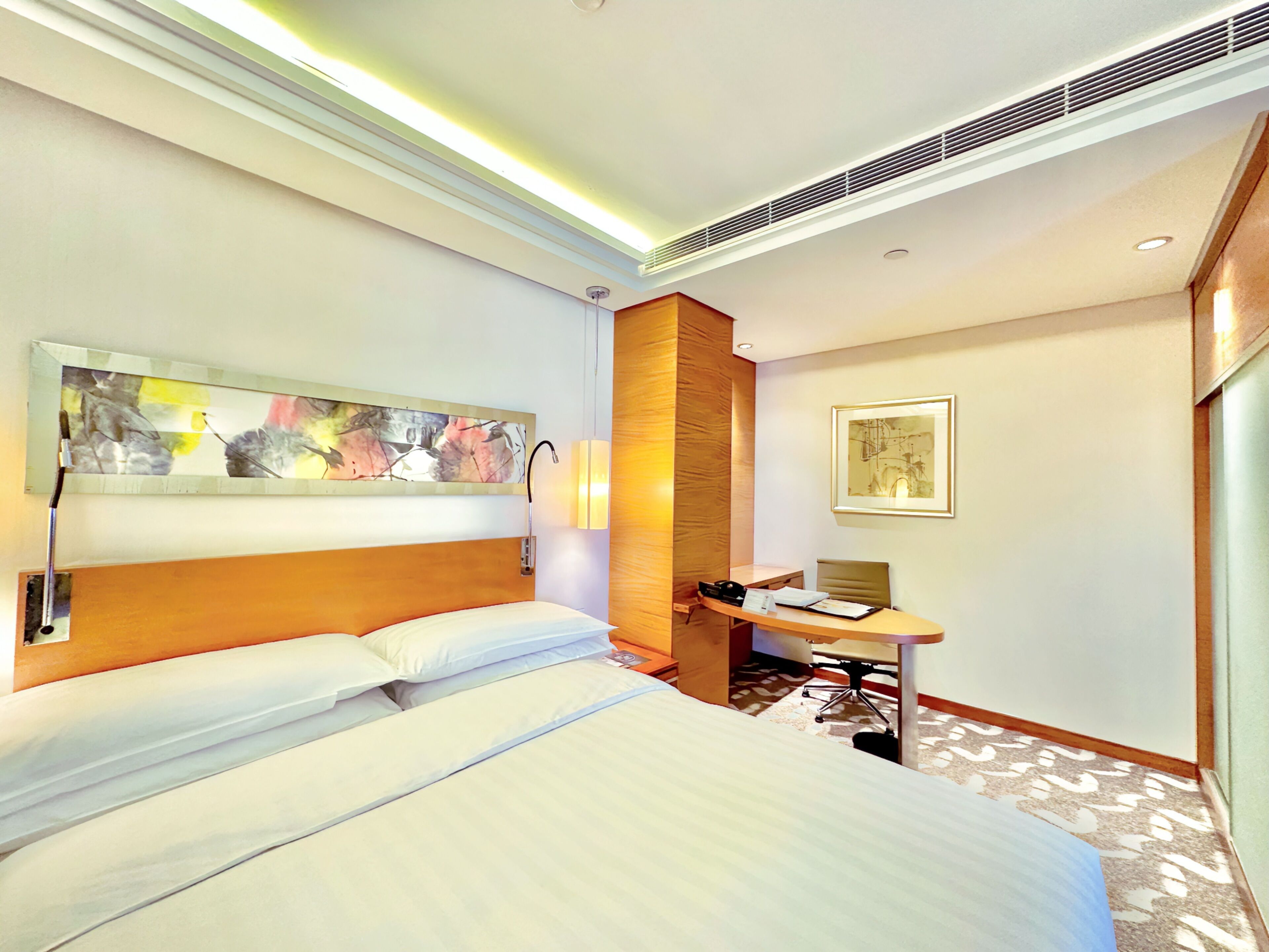 A hotel room with a large bed, a desk with a chair, a wall-mounted artwork, and a ceiling vent.