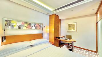 A hotel room with a large bed, a desk with a chair, a wall-mounted artwork, and a ceiling vent.
