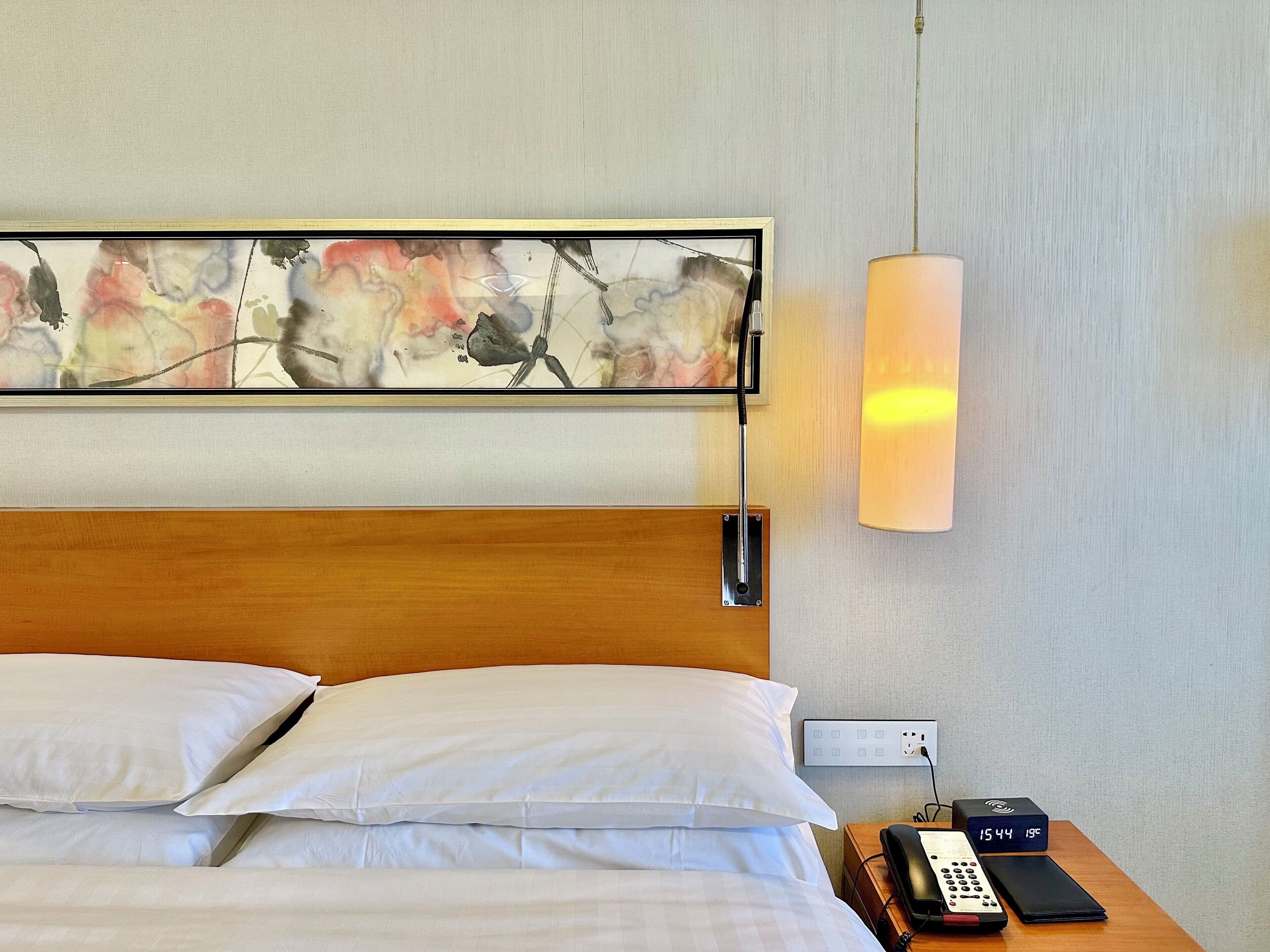 A hotel room with a wooden headboard, white bedding, a wall-mounted abstract painting, a bedside lamp, a power outlet, a digital clock, and a phone on a bedside table.