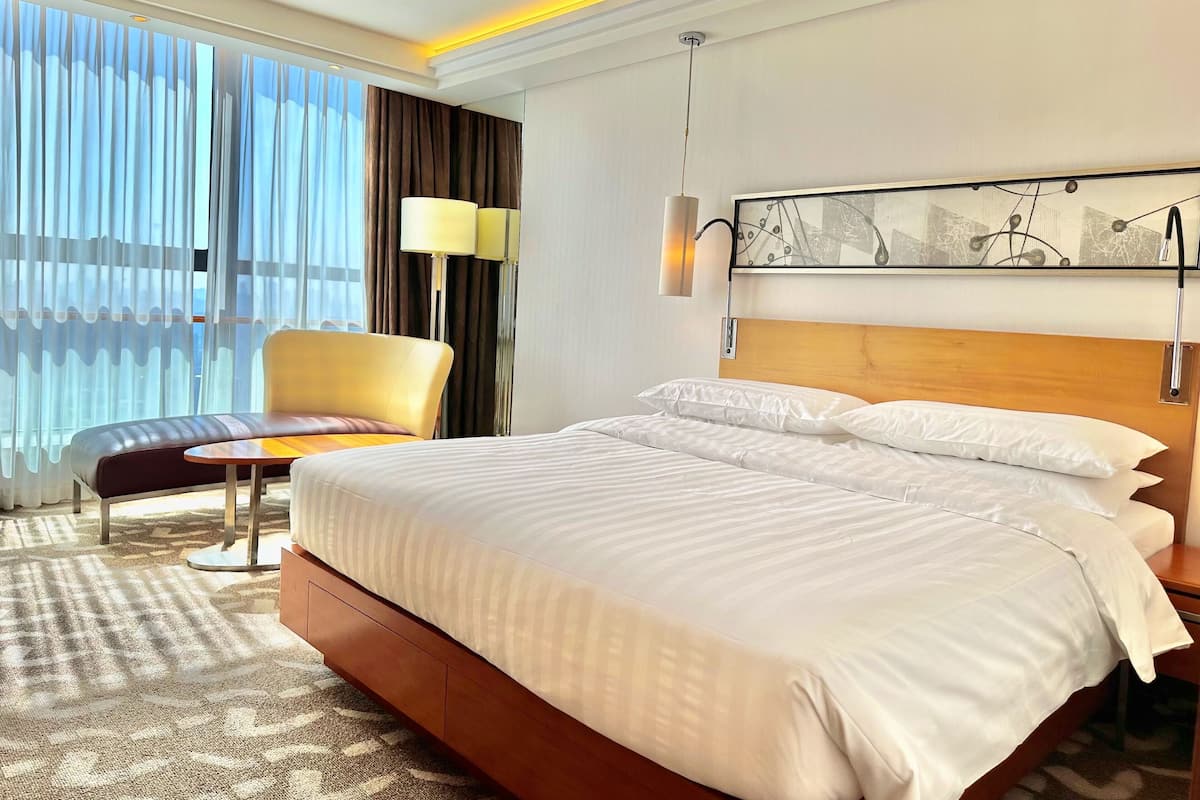 Habitación de hotel con una cama grande, una silla, un escritorio y vistas al exterior a través de cortinas translúcidas.