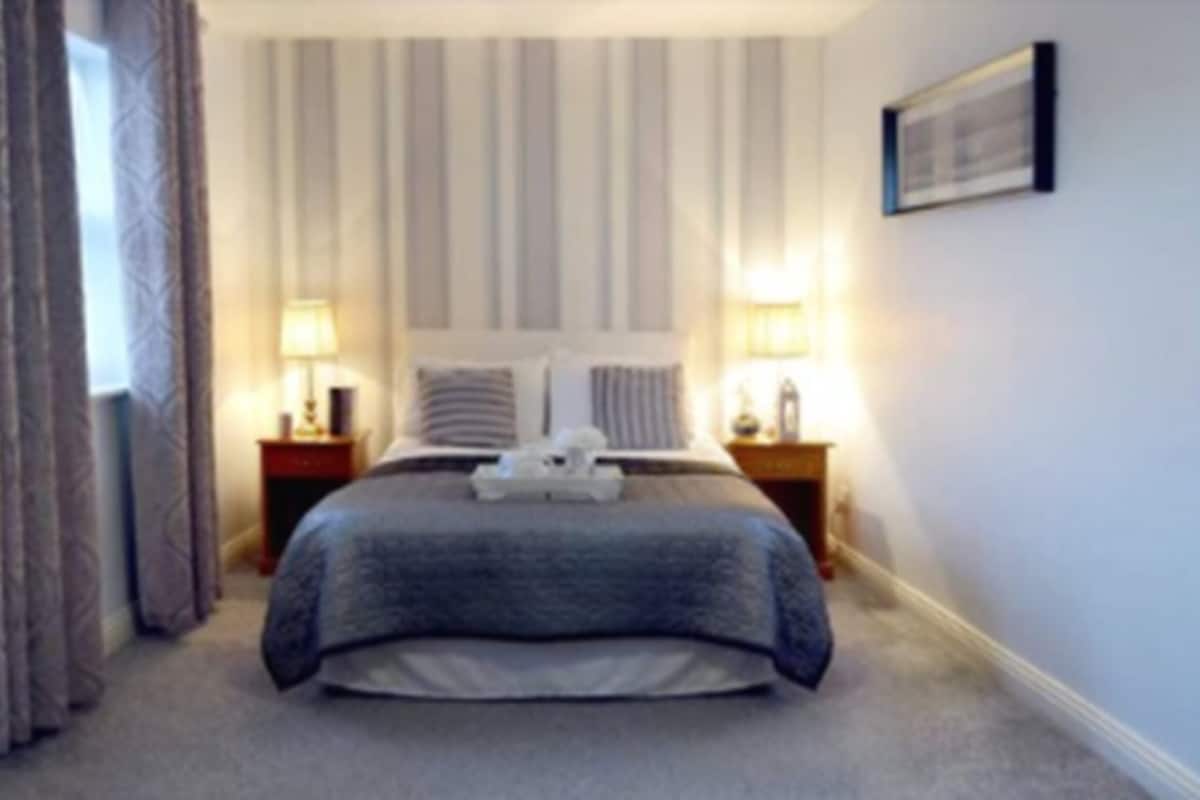 Ein Hotelzimmer mit einem großen Bett, zwei Nachttischen mit Lampen, einem Fenster mit Vorhängen und einem gerahmten Bild an der Wand.