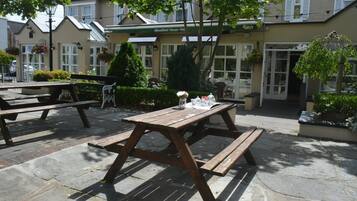 Un área de estar al aire libre con mesas de picnic y bancos de madera, rodeada de follaje y un edificio con un letrero que dice “Woodfield House Hot”.