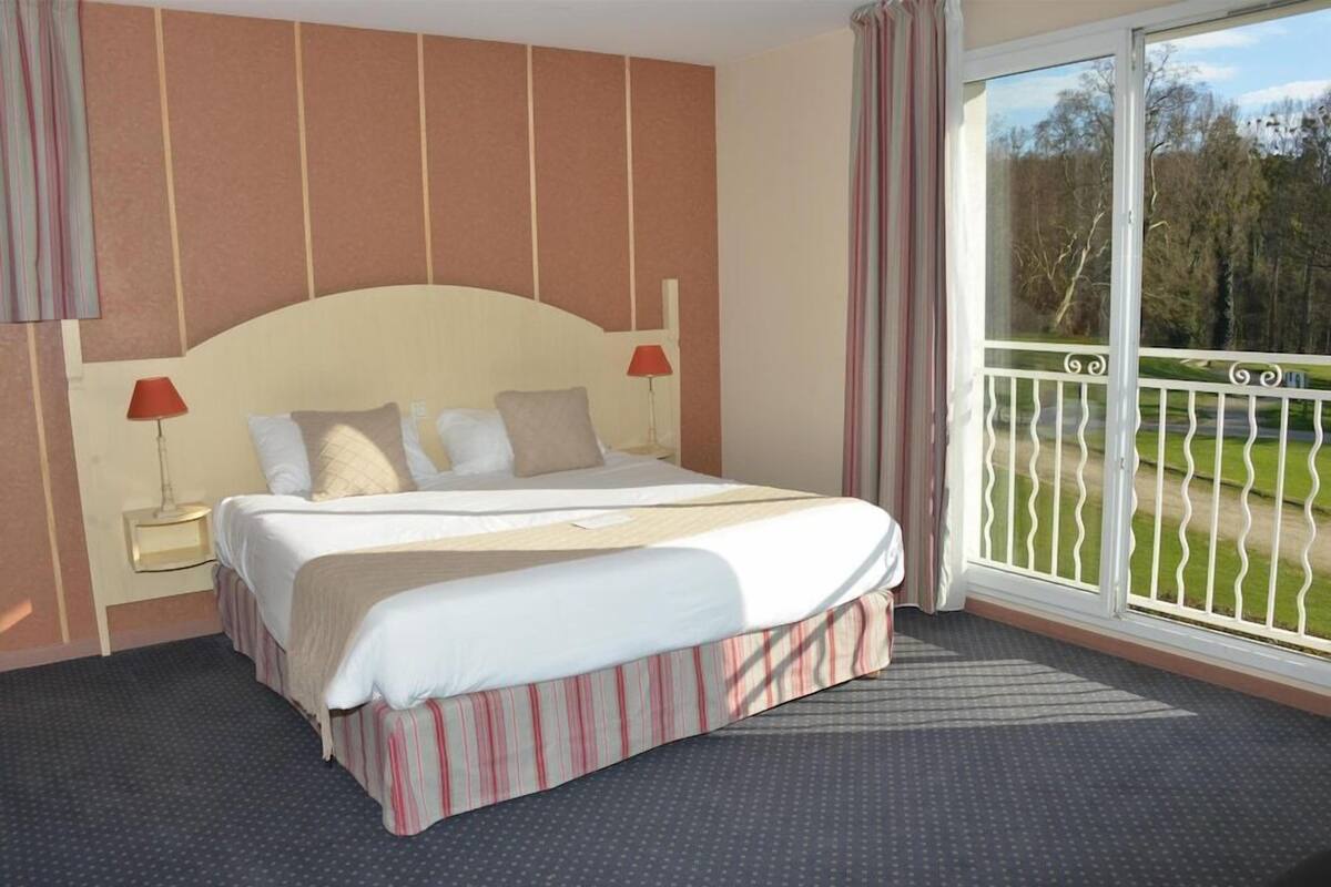 A hotel room with a bed, a nightstand, a lamp, a window with a view of a balcony, and a door.