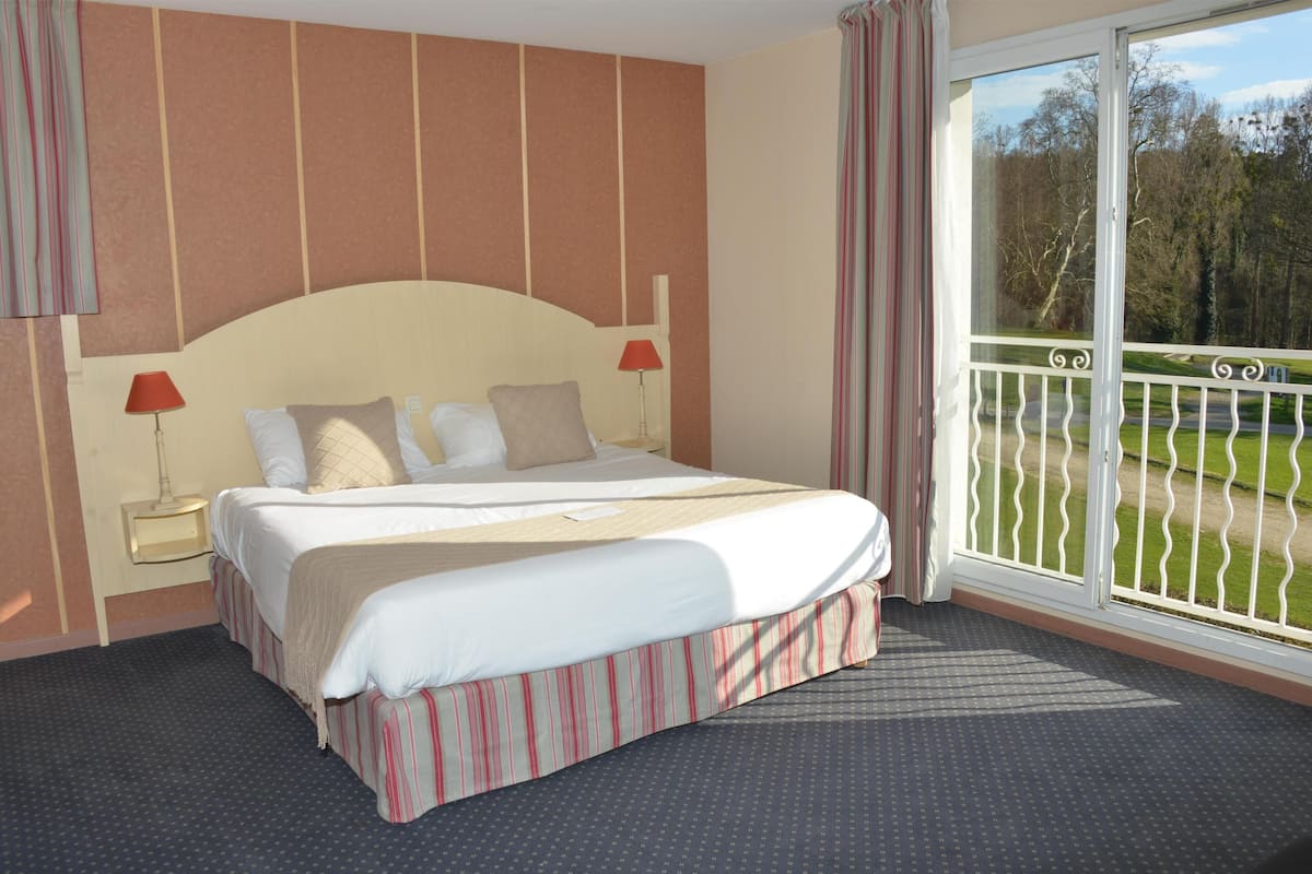 A hotel room with a bed, a nightstand, a lamp, a window with a view of a green landscape, and a balcony with a railing.