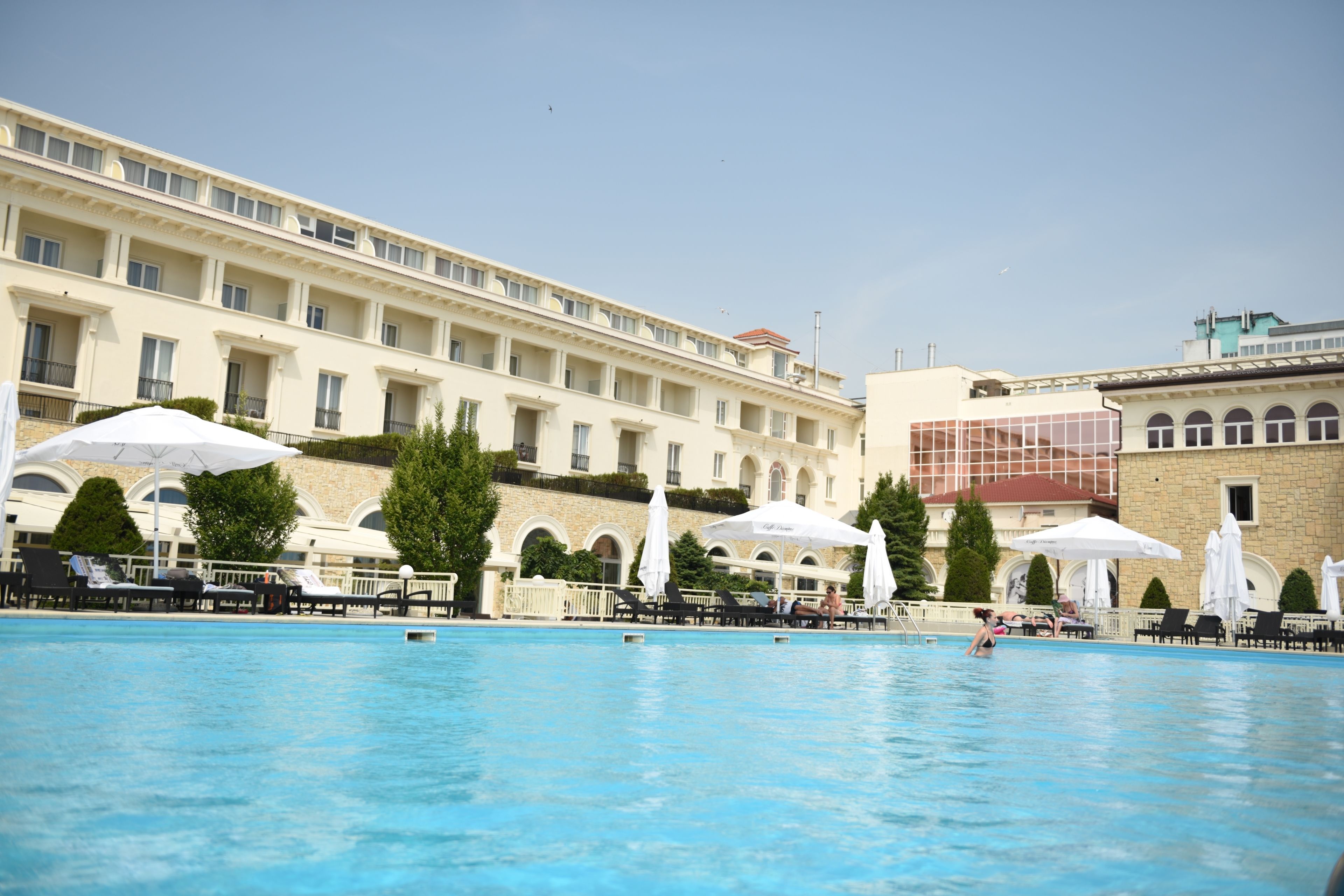 A swimming pool with lounge chairs and umbrellas, surrounded by a building with multiple balconies and arched windows.