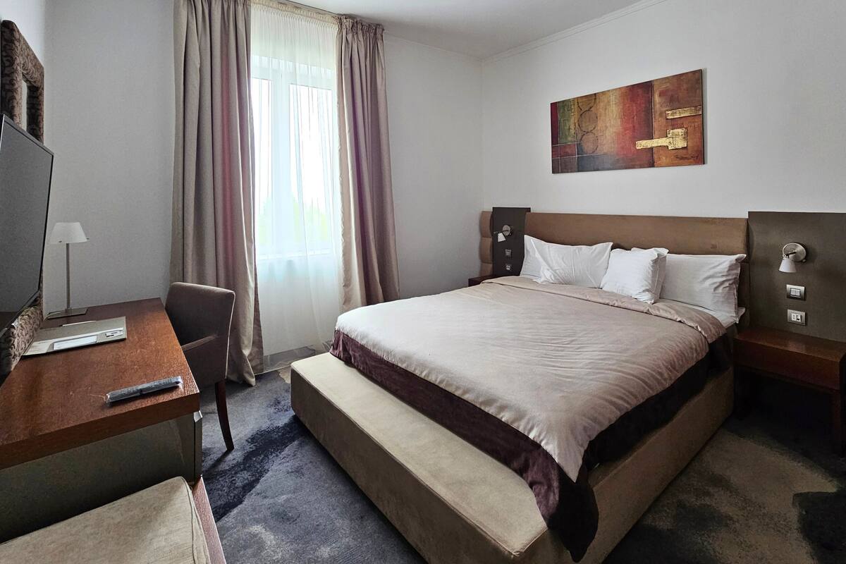 A hotel room with a bed, a desk, a chair, a lamp, and a painting on the wall.