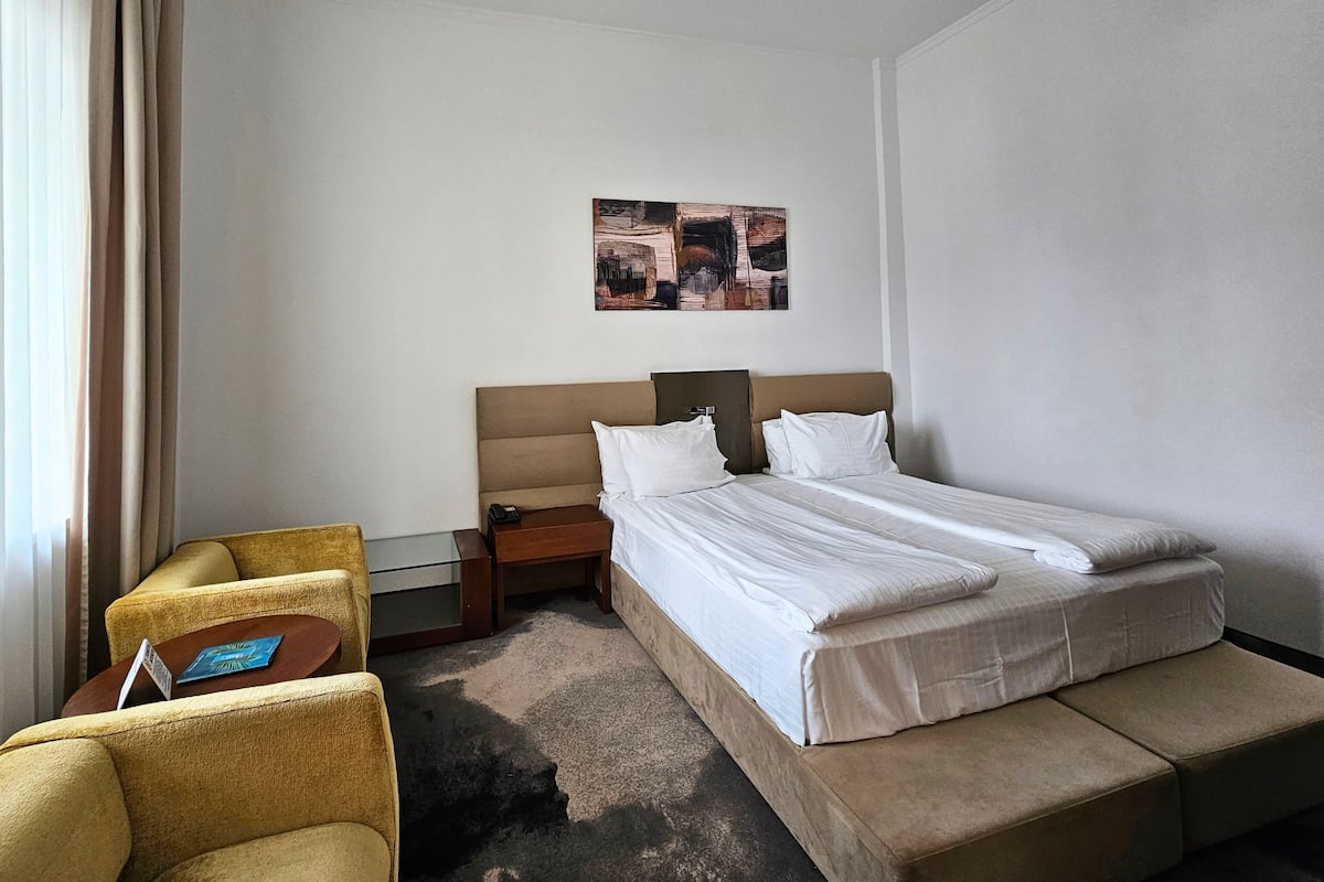 A hotel room with a bed, a bedside table, a chair, and a small table with a book.