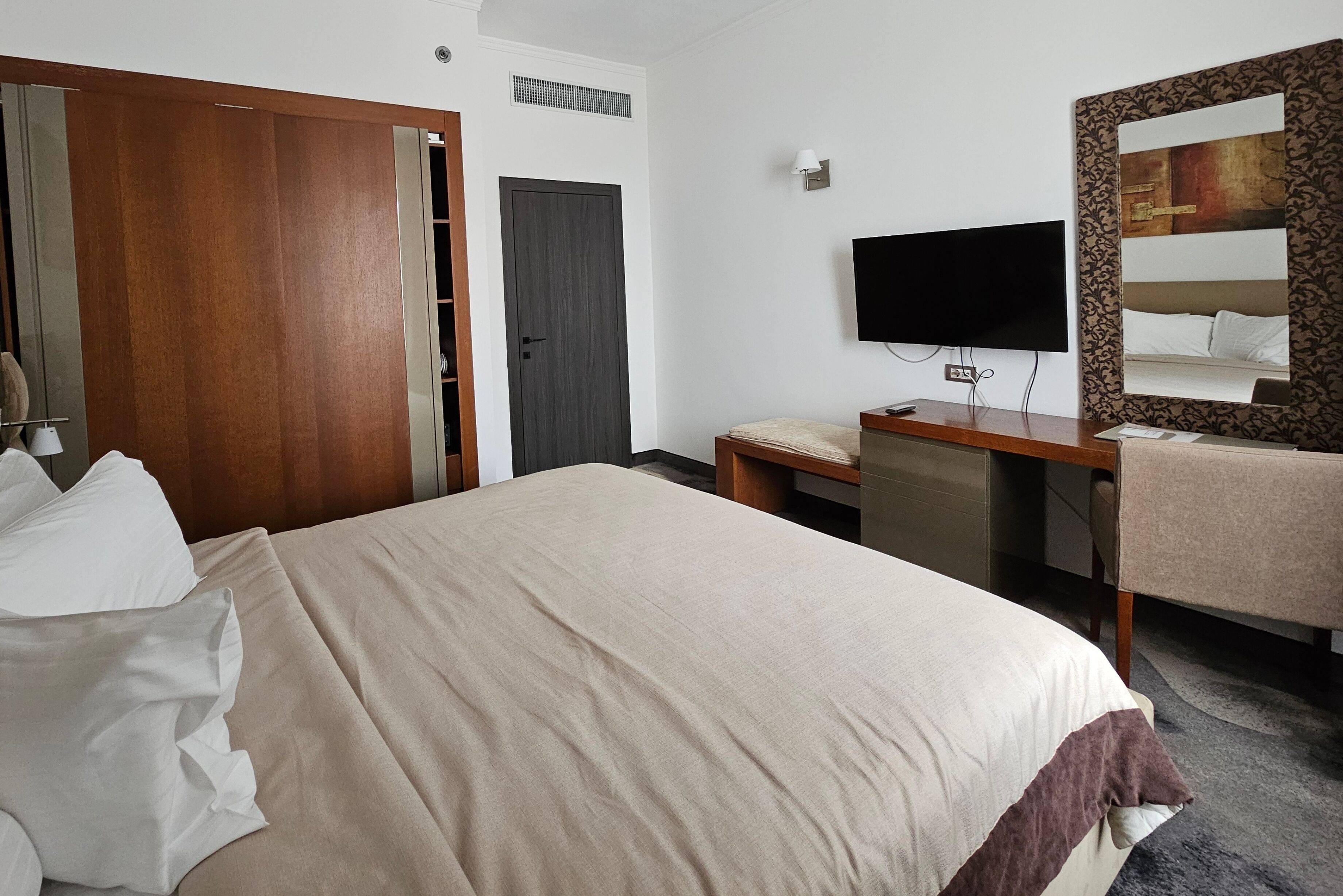 A hotel room with a bed, a desk, a chair, a television, a mirror, and a wardrobe.