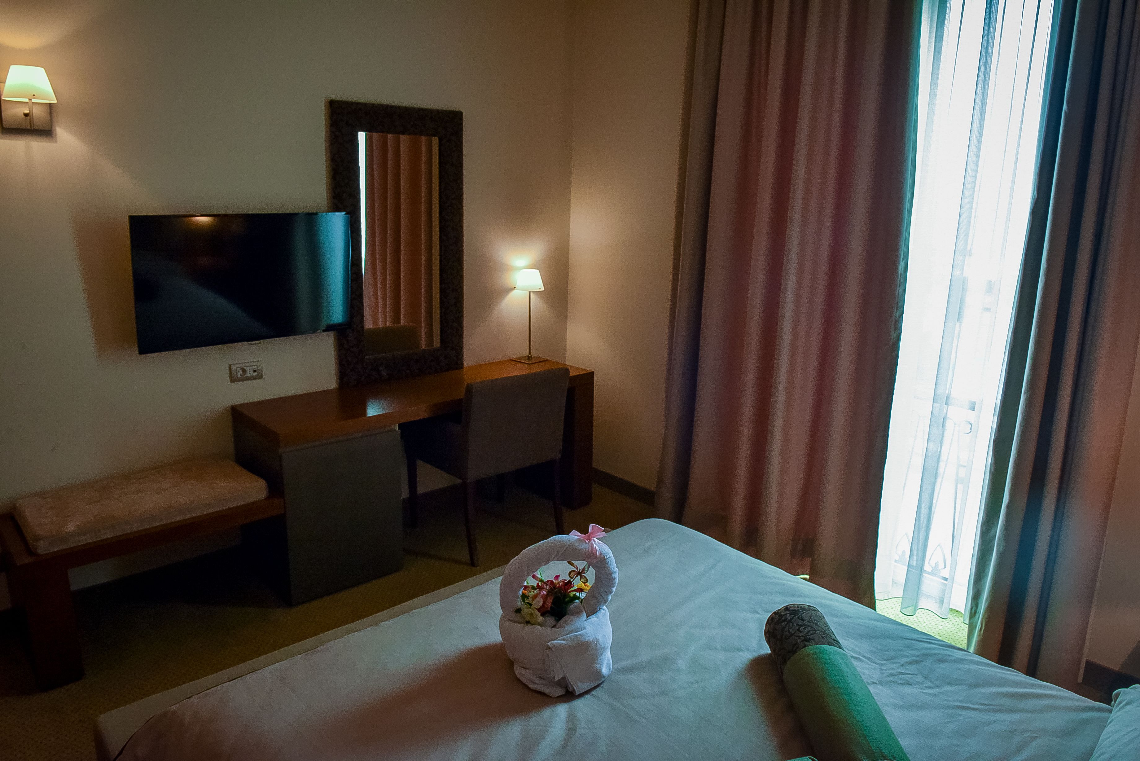 Una camera d'albergo con un letto, una scrivania, una televisione, una lampada e una finestra con tende.