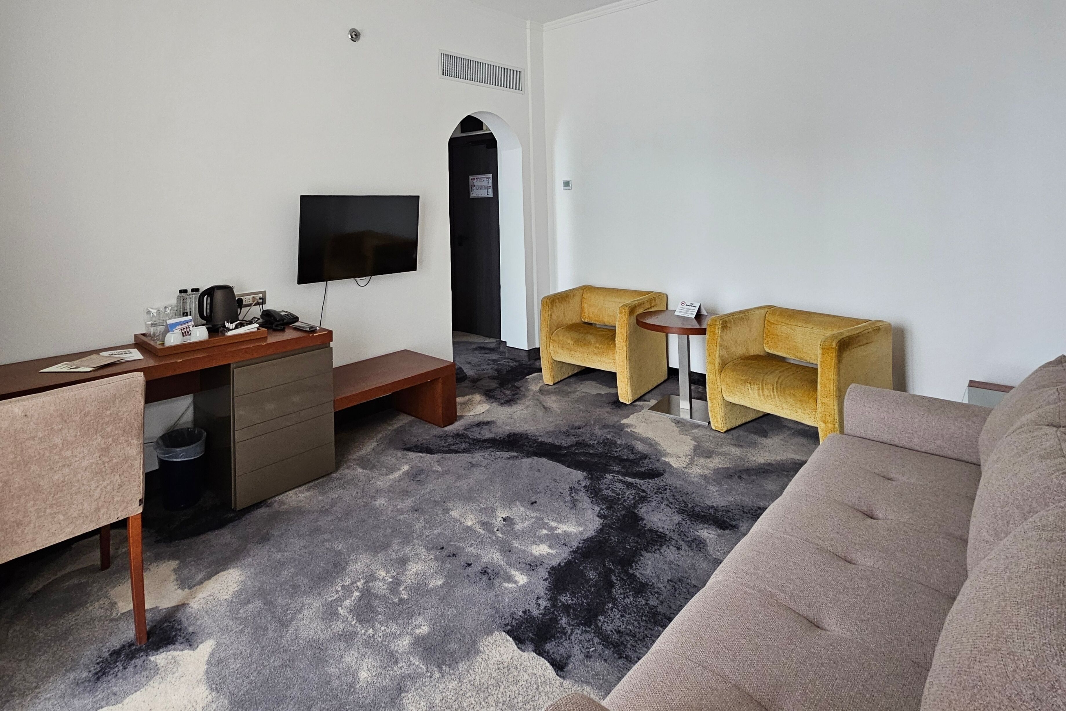A room with a desk, two armchairs, a sofa, a flat-screen TV, and a black and white patterned carpet.