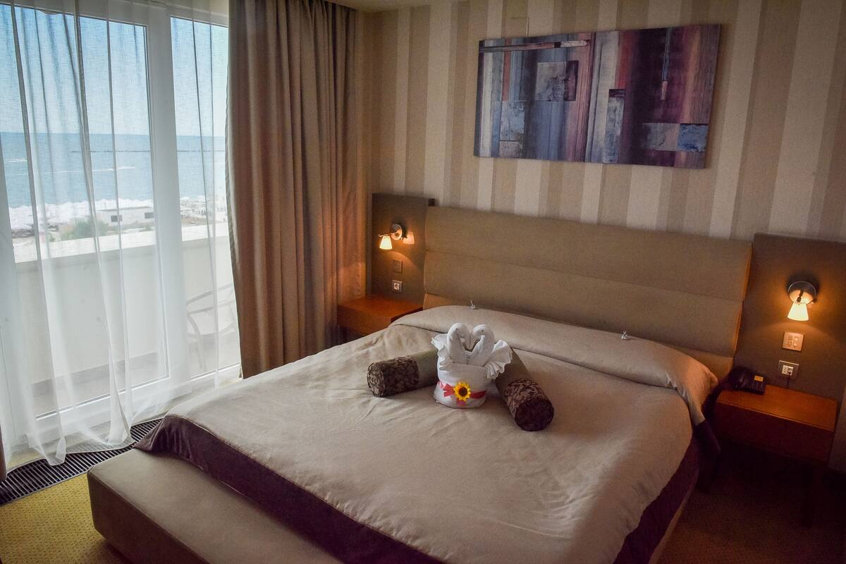 A hotel room with a large bed, a view of the sea, and a painting on the wall.