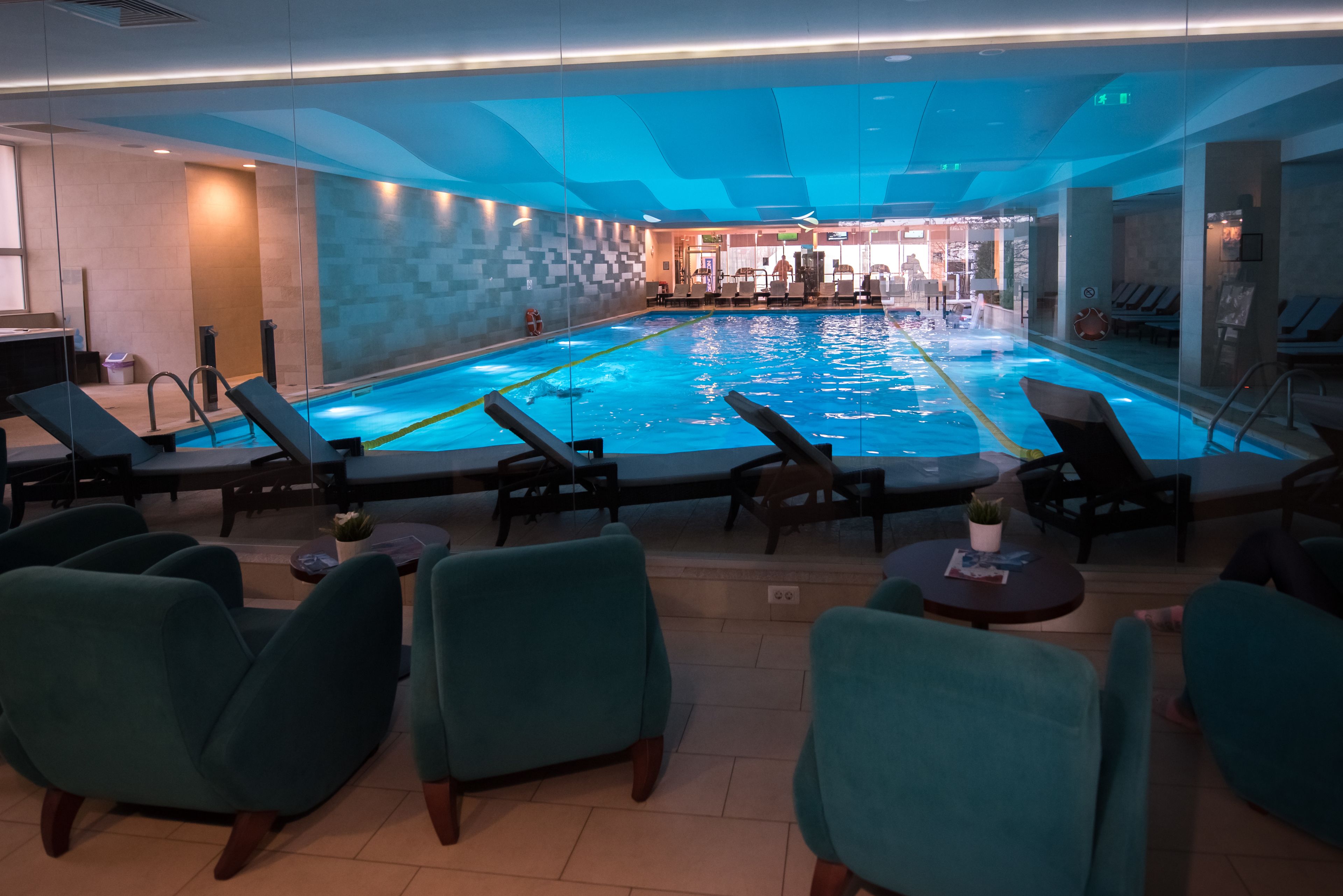 A swimming pool with lounge chairs and tables, surrounded by glass walls and a tiled floor.