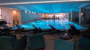A swimming pool with lounge chairs and tables, surrounded by glass walls and a tiled floor.