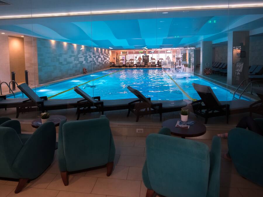 A swimming pool with lounge chairs and tables, surrounded by glass walls and a tiled floor.
