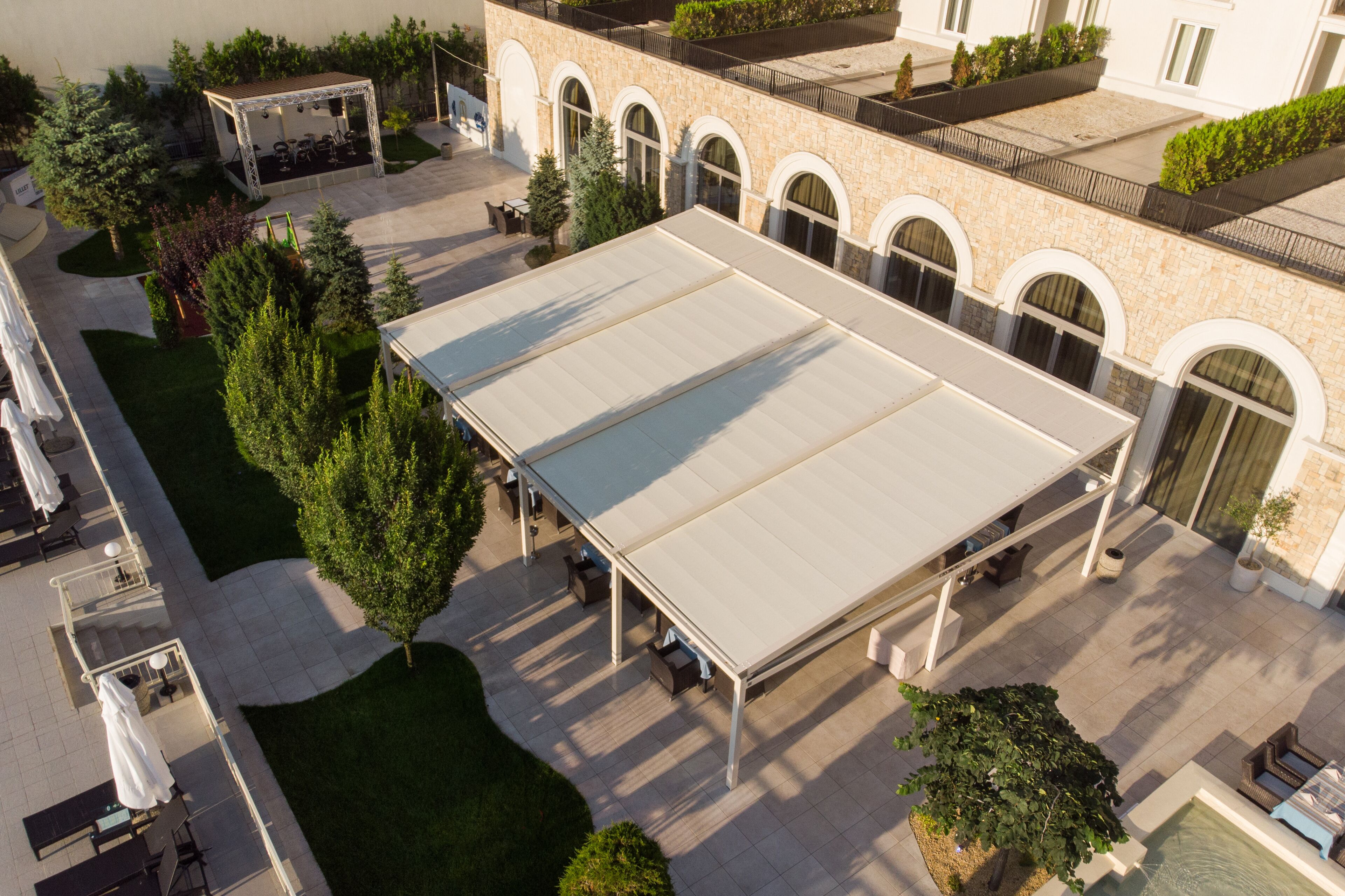 Un espacio de patio al aire libre con zona de estar cubierta, rodeado de árboles y un edificio con ventanas arqueadas.