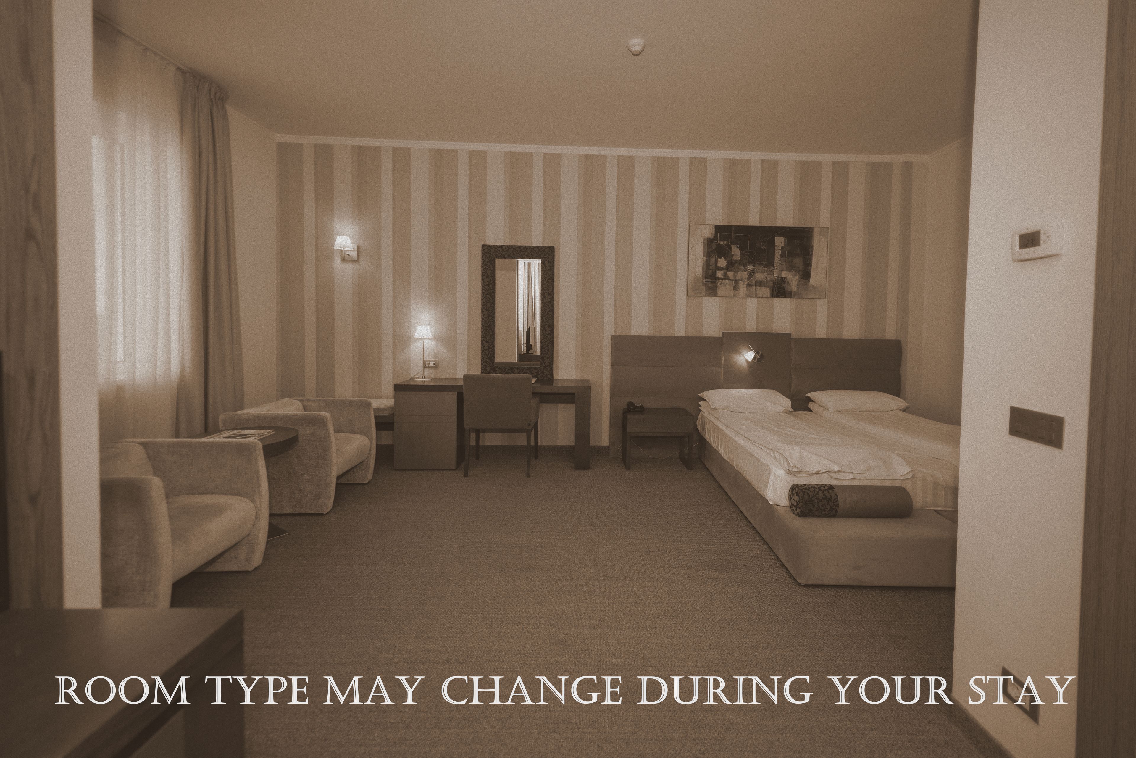 Habitación de hotel con cama, escritorio, silla y sofá. La habitación tiene papel tapiz a rayas y una lámpara de techo.