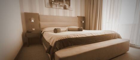 Una habitación de hotel con una cama grande, una mesita de noche, una lámpara de pared y un ventanal con cortinas.