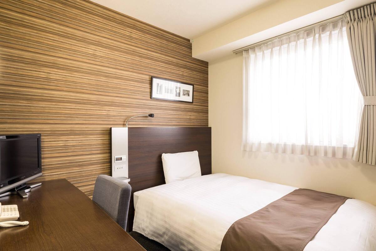 Habitación de hotel con panel de pared de madera, cama con colcha blanca y marrón, escritorio con televisor y teléfono, y ventana con cortinas.